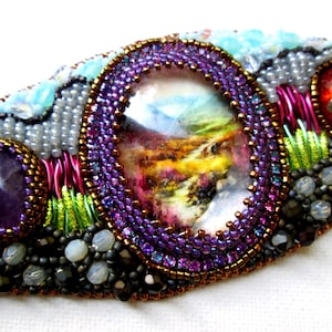 May include: A beaded bracelet with a purple amethyst stone, a red glass bead with a silver dragonfly, and a framed landscape image. The bracelet is made with a variety of beads, including purple, green, and brown.