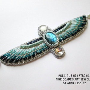 May include: A beaded art jewelry piece in the shape of wings, featuring a central oval blue gemstone and various shades of blue and teal beads. The wings are outlined with silver beads and have a pearl accent at each end. The text "PRECIOUS HEARTBEADS" is visible.
