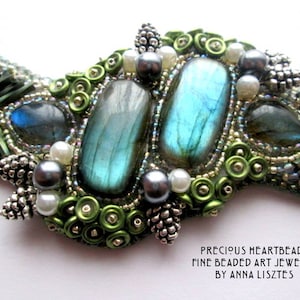 May include: Close-up of a beaded art jewelry piece. The design features large, oval, blue-green labradorite stones, surrounded by green and silver beads, white and gray pearls, and silver accents. The text "PRECIOUS HEARTBEADS FINE BEADED ART JEWELRY BY ANNA LISZTES" is visible.
