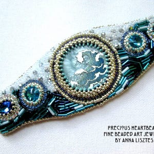 May include: A beaded bracelet with a blue and white wave design. The bracelet is made with small beads and features a large, round, beaded centerpiece. The text "PRECIOUS HEARTBEADS FINE BEADED ART JEWELRY BY ANNA LISZTÉS" is printed below the bracelet.