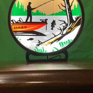 May include: A round, black framed decorative piece featuring a fishing scene. The image shows a person fishing from an orange boat with the word "BASS" on it. A fish is caught on the line, with trees and a lake in the background.
