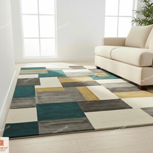 May include: A rectangular area rug with a geometric pattern in shades of teal, gray, cream, and gold. The rug is placed on a light-colored wooden floor, with a cream-colored sofa in the background. The rug's design features large, irregular blocks.