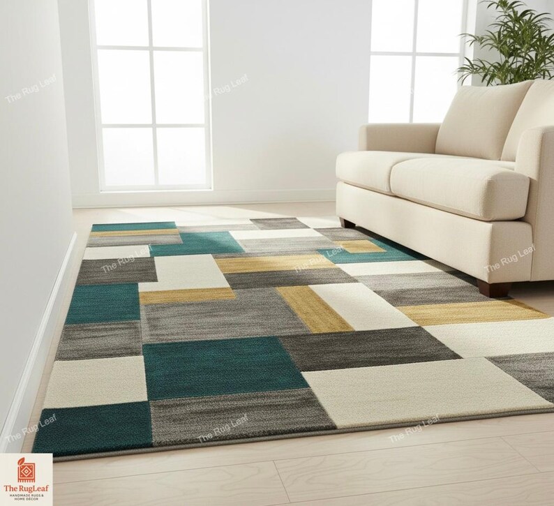 May include: A rectangular area rug with a geometric pattern in shades of teal, gray, cream, and gold. The rug is placed on a light-colored wooden floor, with a cream-colored sofa in the background. The rug's design features large, irregular blocks.
