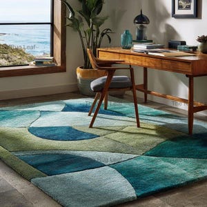 May include: A colourful geometric rug in shades of blue and green. The rug's design features overlapping shapes in teal, seafoam, and deep blue. The rug is in a room with a wooden desk, chair, and a view of the sea through a window.