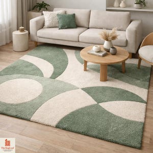 May include: A rectangular area rug featuring a geometric pattern of large, overlapping sage green and cream-coloured circles and arcs. The rug has a thick, textured pile and covers a portion of a light-coloured wooden floor in a living room setting.