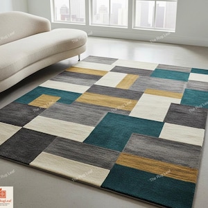 May include: Geometric patterned area rug in shades of teal, gray, gold, and cream. The rug features a modern design with rectangular and square shapes. The rug is placed on a light-colored floor near a beige sofa.