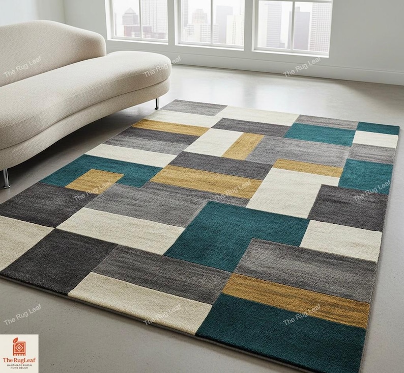 May include: Geometric patterned area rug in shades of teal, gray, gold, and cream. The rug features a modern design with rectangular and square shapes. The rug is placed on a light-colored floor near a beige sofa.