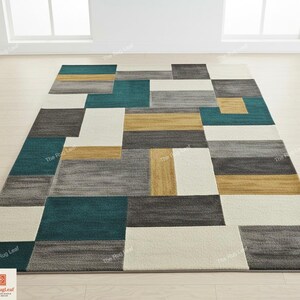 May include: Geometric area rug with a modern design. The rug features rectangular blocks in teal, gray, gold, and off-white. The pattern is a mix of solid and textured sections, creating a contemporary look. The rug is placed on a light wood floor.