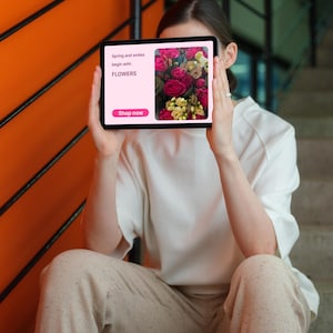 May include: A person holding a tablet displaying a floral arrangement. The tablet screen shows a bouquet of red and yellow roses with the text "Spring and smiles begin with FLOWERS" and a "Shop now" button.