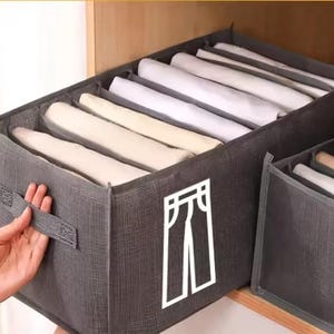 Peut inclure: Organisateur de placard en tissu gris avec plusieurs compartiments, contenant des vêtements blancs et beiges pliés. L'organisateur a une poignée et un contour blanc d'un pantalon sur le devant. L'organisateur est dans une armoire en bois.