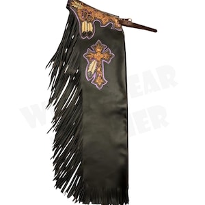 Puede incluir: Chaps de cuero negro con largas franjas laterales. Los chaps presentan bordados decorativos en dorado, morado y marrón claro, incluido un diseño de cruz. Se ve una correa de cuero marrón en la parte superior.