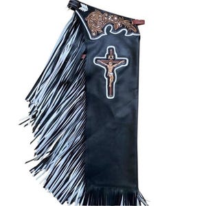 May include: Black leather chaps with white and black fringe. The chaps feature a brown and tan decorative top and a religious cross design. The chaps are designed for riding.