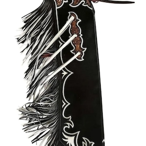 以下が含まれることがあります： 白の装飾トリムとフリンジが付いた黒革のチャップス。チャップスは、茶色のツールレザーのアクセントと白いネクタイが特徴です。ウェスタンスタイルの衣服。