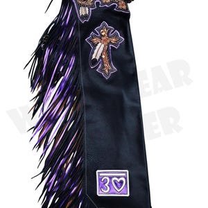 Puede incluir: Chaps de cuero negro con flecos morados y marrones. Los chaps presentan una cruz decorativa y un diseño floral con un parche que dice "30" con un corazón. Los chaps tienen un cinturón de cuero.
