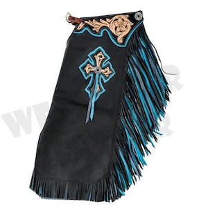 May include: Black leather chaps with turquoise and tan accents. The chaps feature a cross design with a silver-colored center and turquoise fringe along the sides. The top has a floral design and a leather strap.