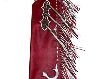 Op de afbeelding: Bordeaux leren chaps met witte en zilveren decoratieve accenten en franjes. De chaps hebben een gedetailleerd bloemmotief op de juk en decoratieve zilveren accenten. De franjes zijn rood, wit en zilver.