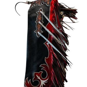 Puede incluir: Chaps de cuero negro y rojo con costuras decorativas y flecos. Los chaps presentan un diseño floral detallado en marrón y blanco, con detalles en rojo y largos flecos en el lateral.