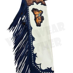 May include: Leather chaps with a white base, navy blue fringe, and decorative stitching. The top features a brown tooled leather design and a cartoon image of a person riding a bull. A brown leather belt is attached.