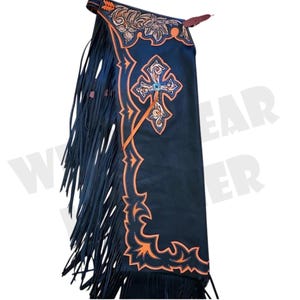 Puede incluir: Chaps de cuero negro con detalles decorativos en naranja y marrón claro. Los chaps presentan un diseño de cruz con una gema azul, patrones florales y geométricos, y largas flecos en los laterales. Prenda de estilo western.