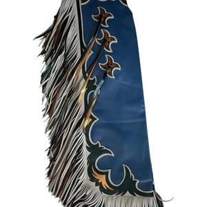 Puede incluir: Chaps de cuero azul con flecos blancos, marrones y negros. Los chaps tienen detalles decorativos dorados y marrones, incluyendo diseños en forma de estrella y un borde festoneado. La parte superior presenta un diseño decorativo marrón y dorado.
