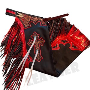 Puede incluir: Chaps de cuero negro con flecos rojos y diseños florales y de cruz decorativos. Los chaps cuentan con una cinturilla roja y negra y lazos de cuero. Los chaps están diseñados con un tema occidental.