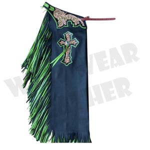 May include: Black leather chaps with neon green fringe and accents. The chaps feature a cross design with a rhinestone center and decorative tooling. A brown leather strap is attached to the chaps.
