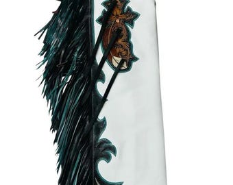 Op de afbeelding: Witte leren chaps met gedetailleerde bruine en teal tooling en franjes. De chaps hebben een decoratief kruisontwerp en zijn afgewerkt met zwart leer en franjes. Een zwarte riem is aan de bovenkant bevestigd.