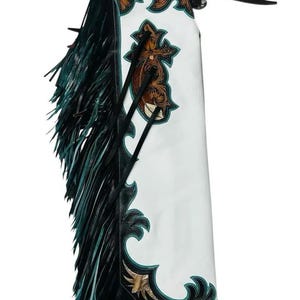 Puede incluir: Chaps de cuero blanco con intrincados detalles en marrón y verde azulado y flecos. Los chaps presentan un diseño de cruz decorativo y están acentuados con ribetes y flecos de cuero negro. Una correa negra está adjunta en la parte superior.