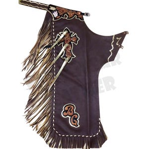 May include: Brown leather chaps with decorative fringe and cross design. Features a belt, decorative stitching, and the initials "BG" in a stylized font. A western-style garment.