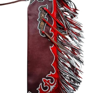 Puede incluir: Chaps de cuero burdeos con intrincados detalles tallados y flecos rojos. Los chaps presentan un diseño decorativo con detalles en blanco y correas de cuero marrón. Los flecos son rojos, blancos y negros.
