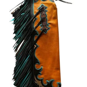 Puede incluir: Chaps de cuero de estilo western en un tono naranja cálido, con intrincados detalles marrones y detalles turquesa. Los chaps tienen largas franjas negras con puntas turquesas y correas negras. Una prenda clásica de rodeo.