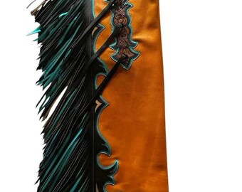 Op de afbeelding: Leren chaps in Western-stijl, in een warme oranje tint, met ingewikkelde bruine details en turquoise accenten. De chaps hebben lange zwarte franjes met turquoise uiteinden en zwarte riemen. Een klassiek rodeo-item.