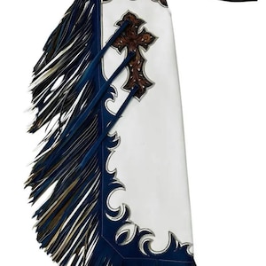 Puede incluir: Chaps de cuero blanco con ribete y flecos azul marino. Los chaps presentan un diseño de cruz marrón y costuras decorativas. Una correa negra se extiende desde la parte superior.