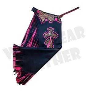 May include: Black and pink western chaps with a cross design. The chaps feature a leather strap, pink fringe, and gold floral accents. The cross is embellished with a silver-colored button.