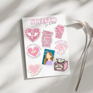 Può includere: Un foglio di adesivi rosa e bianchi con testo in francese. Gli adesivi presentano cuori, un cuore spezzato con un cerotto, un francobollo con una donna e una scatola di fazzoletti. La parola "STYLETO" è in alto.