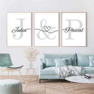 May include: Three framed wall art prints with a minimalist design. Each print features a large initial letter in gray, with a name written in cursive below. The middle print has an ampersand with a heart. The names are Julia and Pascal.