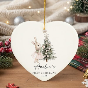 May include: Heart-shaped ceramic Christmas ornament with a bunny decorating a Christmas tree. The ornament has a gold string for hanging and features the text "Amelia's First Christmas 2025". Festive holiday decor.