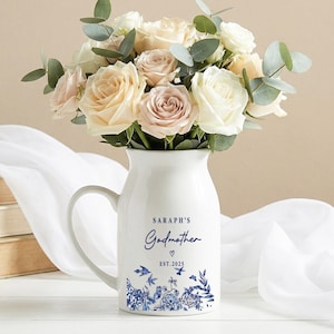 Puede incluir: Una jarra de cerámica blanca con asa, llena de un ramo de rosas blancas, rosas y melocotón, y hojas de eucalipto. La jarra está decorada con diseños florales azules y el texto "SARAPH'S Godmother EST. 2025".