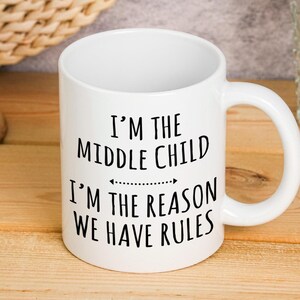 Könnte beinhalten: Weiße Keramik-Tasse mit dem Text "I'M THE MIDDLE CHILD I'M THE REASON WE HAVE RULES" in Schwarz. Die Tasse hat einen gebogenen Henkel und steht auf einer Holzoberfläche.