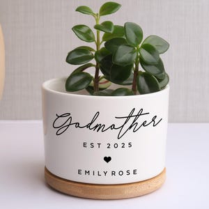 Pode incluir: Um vaso de cerâmica branco com a palavra "Godmother" em escrita cursiva, "EST 2025", um coração e "EMILY ROSE" impressos em preto. O vaso contém uma pequena planta verde e repousa sobre uma base de madeira.
