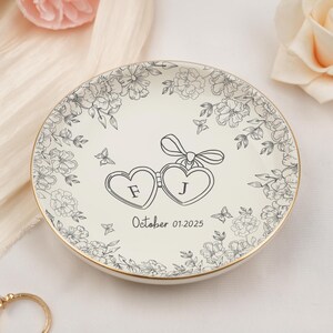 May include: A white ceramic ring dish with a gold rim, featuring a floral design and two heart-shaped frames with the letters "F" and "J". The date "October 01.2025" is printed below. Decorative butterflies are also included.