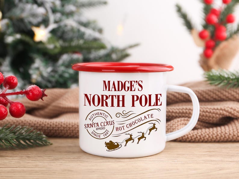 May include: White enamel mug with a red rim and handle. The mug features the text "Madge's North Pole" in a festive font, along with a Santa Claus seal and a hot chocolate graphic. A charming holiday-themed item.