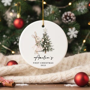 May include: A white ceramic Christmas ornament with a gold string. The ornament features a watercolor illustration of a bunny wearing a scarf and tutu next to a Christmas tree. Text reads "Amelia's First Christmas 2025".