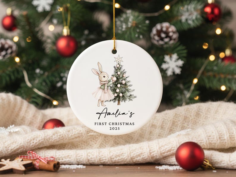 May include: A white ceramic Christmas ornament with a gold string. The ornament features a watercolor illustration of a bunny wearing a scarf and tutu next to a Christmas tree. Text reads "Amelia's First Christmas 2025".