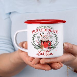 May include: White enamel mug with a red rim and handle, featuring a festive design. The mug has the words "North Pole Hot Chocolate" and a graphic of a hot chocolate cup with whipped cream and a candy cane. The mug is held in two hands.