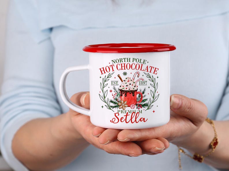 May include: White enamel mug with a red rim and handle, featuring a festive design. The mug has the words "North Pole Hot Chocolate" and a graphic of a hot chocolate cup with whipped cream and a candy cane. The mug is held in two hands.