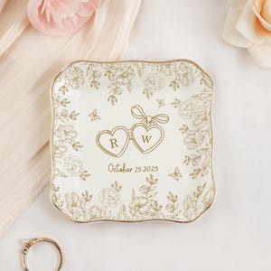 May include: A square, off-white ceramic ring dish with a gold rim and floral design. Two connected heart-shaped pendants with the letters "R" and "W" are in the center, with the date "October 25, 2025" below. A gold ring is in the lower left corner.