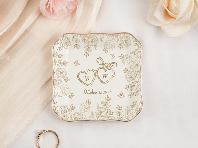 May include: A square, off-white ceramic ring dish with a gold rim and floral design. Two connected heart-shaped pendants with the letters "R" and "W" are in the center, with the date "October 25, 2025" below. A gold ring is in the lower left corner.