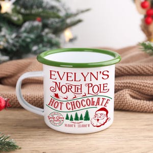 May include: White enamel mug with a green rim and handle, featuring the text "Evelyn's North Pole Hot Chocolate" in red. The design includes Santa Claus, a sleigh, and Christmas trees. Coordinates are printed at the bottom.