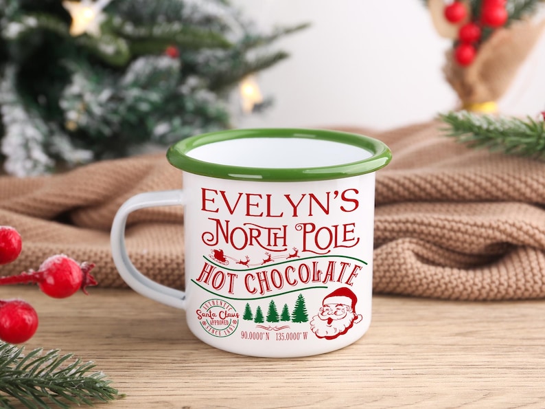 May include: White enamel mug with a green rim and handle, featuring the text "Evelyn's North Pole Hot Chocolate" in red. The design includes Santa Claus, a sleigh, and Christmas trees. Coordinates are printed at the bottom.